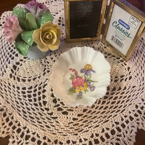 May include: A set of two gold-toned picture frames with a floral porcelain figurine and a small white porcelain dish with a floral design. The frames are both open and the larger frame has the text "2 1/4 x 3 1/4 Photo Frame" printed on the front. The smaller frame has the text "ECKERD Classics School Size Photo Frame" printed on the front. The frames are sitting on a white crocheted doily.