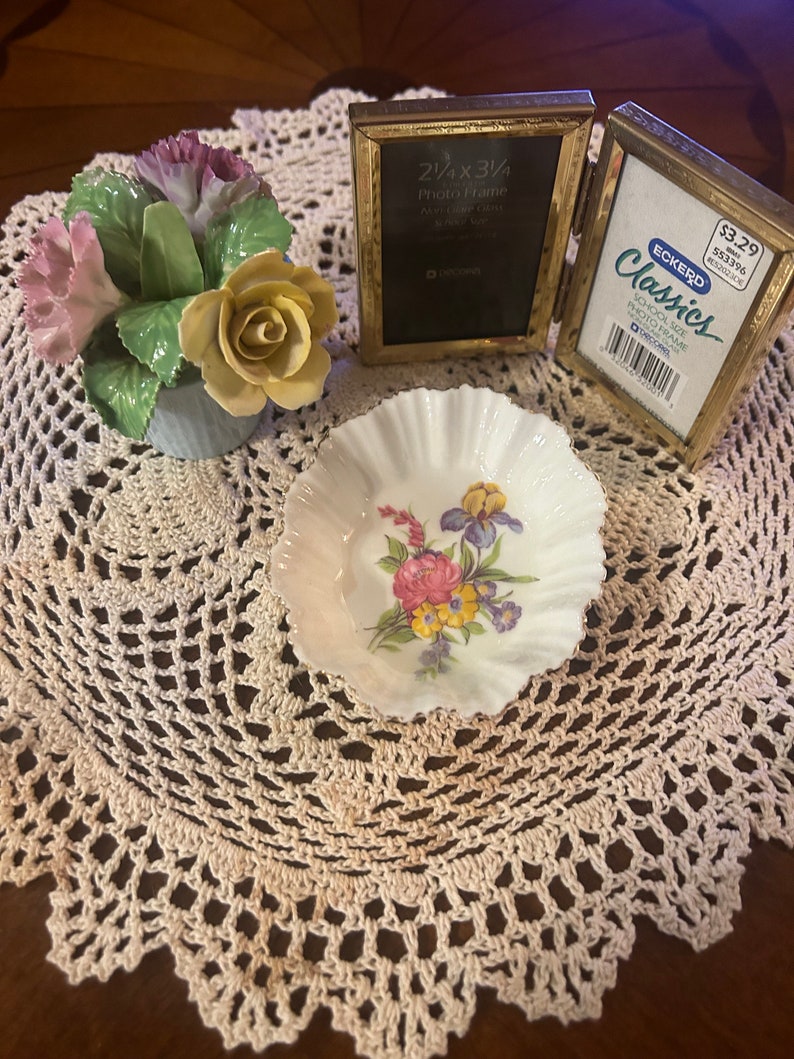 May include: A set of two gold-toned picture frames with a floral porcelain figurine and a small white porcelain dish with a floral design. The frames are both open and the larger frame has the text "2 1/4 x 3 1/4 Photo Frame" printed on the front. The smaller frame has the text "ECKERD Classics School Size Photo Frame" printed on the front. The frames are sitting on a white crocheted doily.