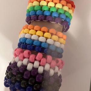 May include: A stack of colorful beaded bracelets in rainbow, white, pink, purple, black, and blue colors. The bracelets are made of small plastic beads and are worn on a person's wrist.