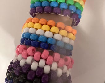 May include: A stack of colorful beaded bracelets in rainbow, white, pink, purple, black, and blue colors. The bracelets are made of small plastic beads and are worn on a person's wrist.