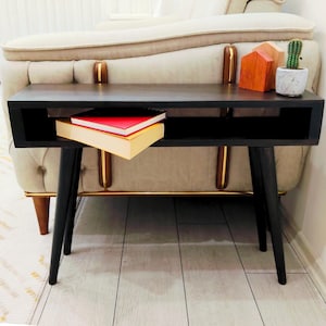 Puede incluir: Una mesa auxiliar negra de estilo moderno de mediados de siglo con patas anguladas. La mesa tiene un estante con dos libros. Un sofá beige con ribetes dorados está en el fondo. Casas de madera decorativas y un pequeño cactus en maceta están sobre la mesa.