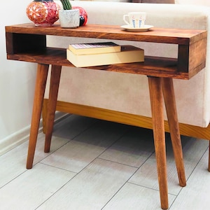 Peut inclure: Une table d'appoint en bois de style moderne du milieu du siècle, avec un plateau rectangulaire et une étagère inférieure. La table a quatre pieds inclinés et est teintée d'une couleur marron chaud. Une tasse et une soucoupe, des livres et des objets décoratifs sont exposés.