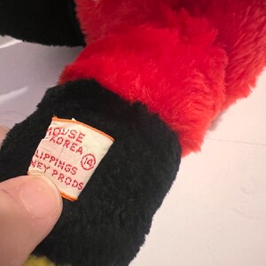 Pode incluir: Close-up de um brinquedo de pel&uacute;cia nas cores vermelho, preto e amarelo. O brinquedo tem uma etiqueta que diz "HOUSE KOREA LIPPINGS DISNEY PRODS". O design do brinquedo lembra um personagem de desenho animado.