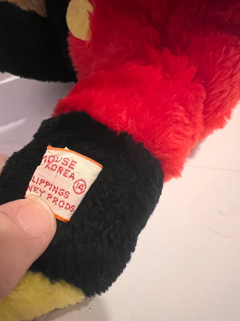 Pode incluir: Close-up de um brinquedo de pel&uacute;cia nas cores vermelho, preto e amarelo. O brinquedo tem uma etiqueta que diz "HOUSE KOREA LIPPINGS DISNEY PRODS". O design do brinquedo lembra um personagem de desenho animado.