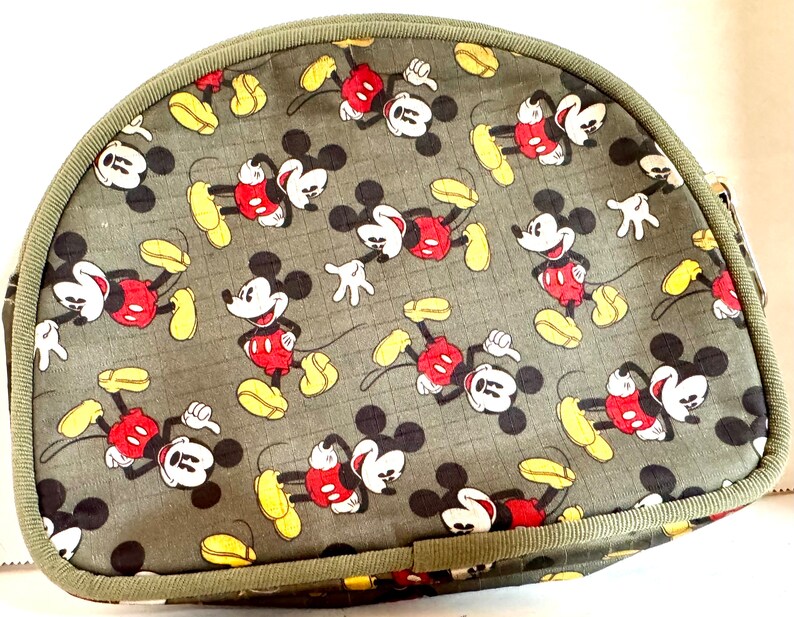 May include: A half-moon shaped bag with a repeating pattern of Mickey Mouse in red shorts, yellow shoes, and white gloves on a gray background. The bag has a green trim and a zipper closure.