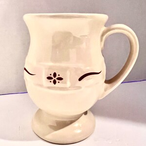 May include: Cream-colored ceramic mug with a whimsical face design. The mug features a rounded base, a curved handle, and a decorative floral accent. The eyes are represented by curved lines.