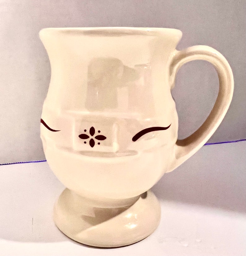 May include: Cream-colored ceramic mug with a whimsical face design. The mug features a rounded base, a curved handle, and a decorative floral accent. The eyes are represented by curved lines.