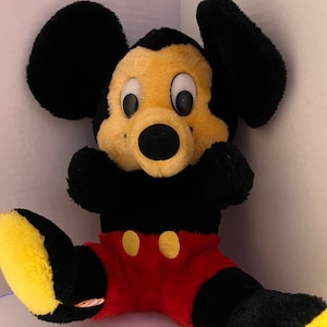Pode incluir: Um brinquedo de pel&uacute;cia do Mickey Mouse com orelhas pretas, corpo preto e cal&ccedil;&otilde;es vermelhos. O brinquedo tem um rosto e p&eacute;s amarelos, com olhos grandes e redondos e um nariz preto. Dois bot&otilde;es amarelos adornam a frente dos cal&ccedil;&otilde;es. O brinquedo est&aacute; sentado.