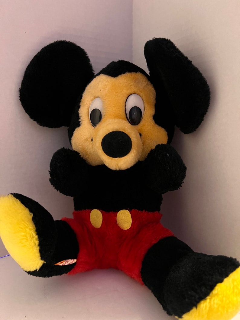 Pode incluir: Um brinquedo de pel&uacute;cia do Mickey Mouse com orelhas pretas, corpo preto e cal&ccedil;&otilde;es vermelhos. O brinquedo tem um rosto e p&eacute;s amarelos, com olhos grandes e redondos e um nariz preto. Dois bot&otilde;es amarelos adornam a frente dos cal&ccedil;&otilde;es. O brinquedo est&aacute; sentado.