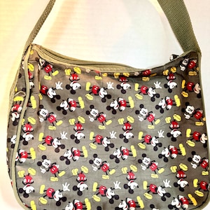 May include: Olive green shoulder bag featuring a repeating pattern of Mickey Mouse in red, black, yellow, and white. The bag has a zippered top and a matching olive green shoulder strap. The bag is made of a lightweight material.
