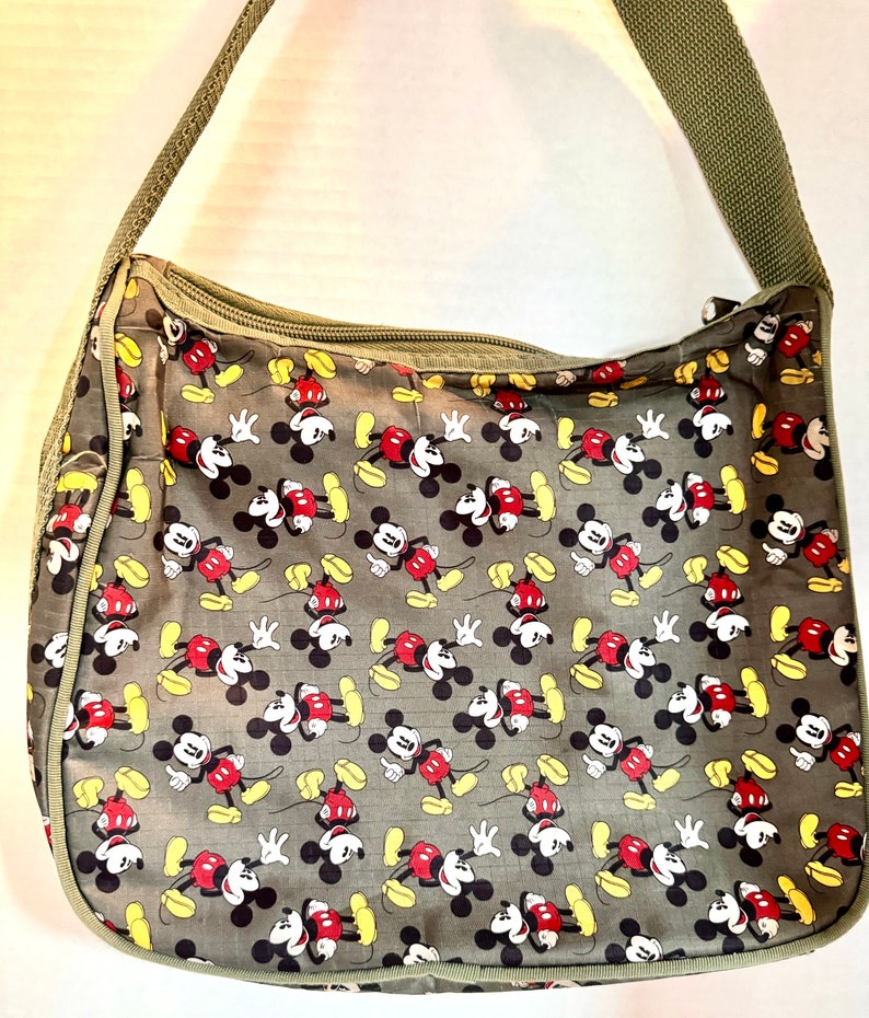 May include: Olive green shoulder bag featuring a repeating pattern of Mickey Mouse in red, black, yellow, and white. The bag has a zippered top and a matching olive green shoulder strap. The bag is made of a lightweight material.