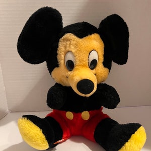 Pode incluir: Um brinquedo de pel&uacute;cia do Mickey Mouse com orelhas, bra&ccedil;os e pernas pretas, rosto amarelo e cal&ccedil;&otilde;es vermelhos. O brinquedo tem olhos grandes e redondos e um nariz preto. Os p&eacute;s s&atilde;o amarelos. O brinquedo est&aacute; sentado com as pernas abertas.