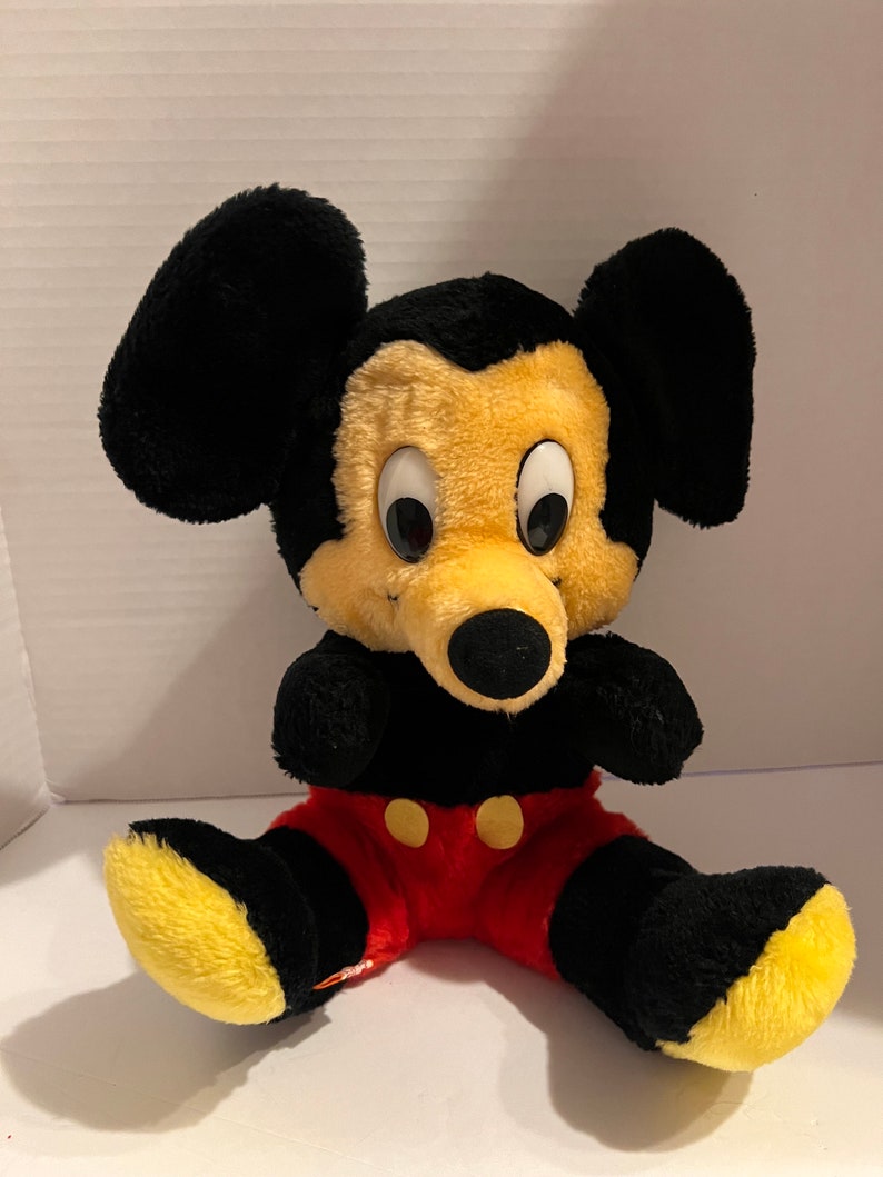 Pode incluir: Um brinquedo de pel&uacute;cia do Mickey Mouse com orelhas, bra&ccedil;os e pernas pretas, rosto amarelo e cal&ccedil;&otilde;es vermelhos. O brinquedo tem olhos grandes e redondos e um nariz preto. Os p&eacute;s s&atilde;o amarelos. O brinquedo est&aacute; sentado com as pernas abertas.