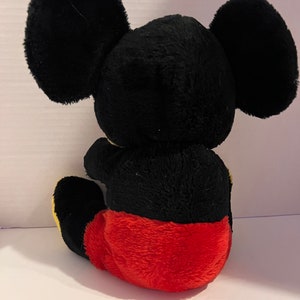 Pode incluir: Um brinquedo de pel&uacute;cia de um rato de desenho animado, visto por tr&aacute;s. O brinquedo tem grandes orelhas pretas, um corpo preto e cal&ccedil;&otilde;es vermelhos. O brinquedo est&aacute; sentado, com os p&eacute;s amarelos vis&iacute;veis.