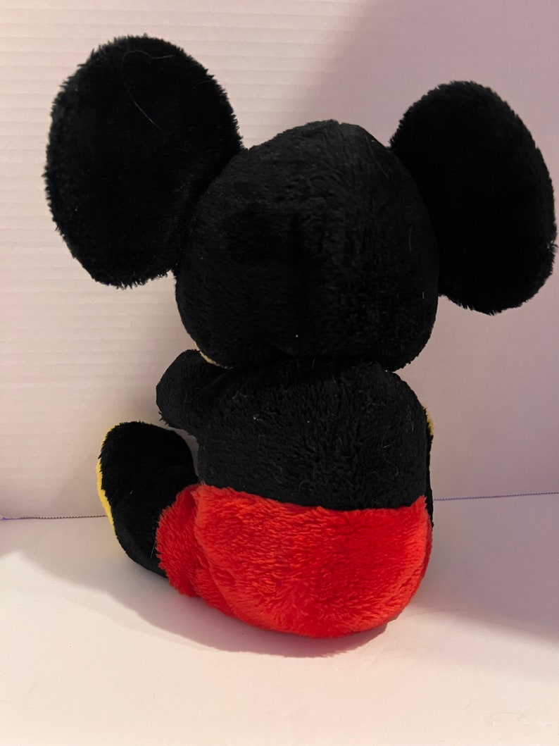 Pode incluir: Um brinquedo de pel&uacute;cia de um rato de desenho animado, visto por tr&aacute;s. O brinquedo tem grandes orelhas pretas, um corpo preto e cal&ccedil;&otilde;es vermelhos. O brinquedo est&aacute; sentado, com os p&eacute;s amarelos vis&iacute;veis.