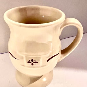 May include: Cream-colored ceramic mug with a rounded base and a curved handle. The mug features a decorative design with two curved lines and a small floral pattern in dark brown. The mug is designed for hot beverages.