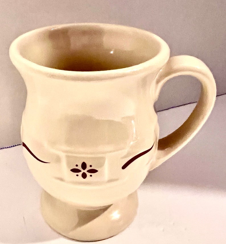 May include: Cream-colored ceramic mug with a rounded base and a curved handle. The mug features a decorative design with two curved lines and a small floral pattern in dark brown. The mug is designed for hot beverages.