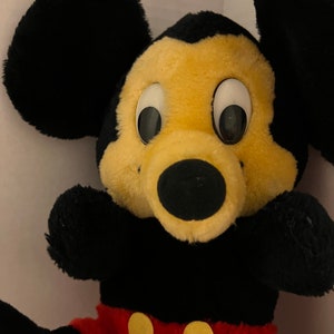 Pode incluir: Um brinquedo de pel&uacute;cia do Mickey Mouse com grandes orelhas pretas, um rosto amarelo e um nariz preto. O brinquedo tem olhos brancos grandes e usa cal&ccedil;&otilde;es vermelhos com bot&otilde;es amarelos. Os bra&ccedil;os e o corpo s&atilde;o pretos.