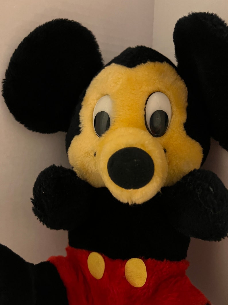 Pode incluir: Um brinquedo de pel&uacute;cia do Mickey Mouse com grandes orelhas pretas, um rosto amarelo e um nariz preto. O brinquedo tem olhos brancos grandes e usa cal&ccedil;&otilde;es vermelhos com bot&otilde;es amarelos. Os bra&ccedil;os e o corpo s&atilde;o pretos.