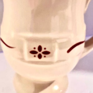 May include: Cream-colored ceramic pitcher with a decorative design. The pitcher features a dark red floral pattern and accents. The handle is visible on the right side. The pitcher has a rounded base and a flared top.