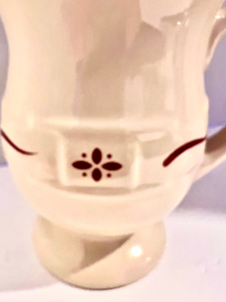 May include: Cream-colored ceramic pitcher with a decorative design. The pitcher features a dark red floral pattern and accents. The handle is visible on the right side. The pitcher has a rounded base and a flared top.