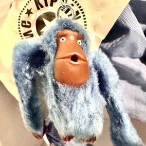 May include: A blue plush monkey keychain with a brown face and open mouth. The keychain has a silver metal ring and is attached to a tag with the brand name "KIP". The monkey has a fluffy texture.