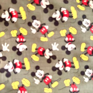 May include: Fabric with a repeating pattern of Mickey Mouse. The cartoon character is depicted in his classic outfit: red shorts, white gloves, and yellow shoes. The background is a muted green color.