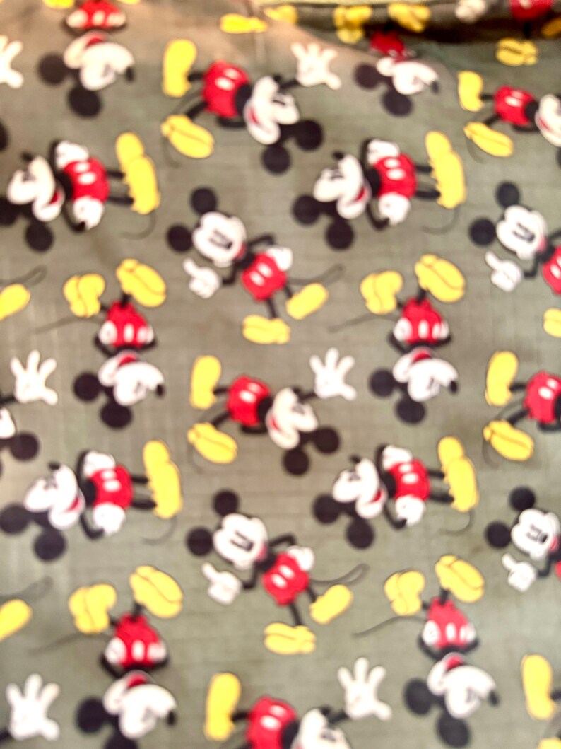 May include: Fabric with a repeating pattern of Mickey Mouse. The cartoon character is depicted in his classic outfit: red shorts, white gloves, and yellow shoes. The background is a muted green color.