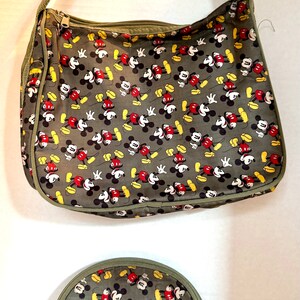 May include: A gray shoulder bag and matching cosmetic bag feature a repeating pattern of Mickey Mouse in red, black, yellow, and white. The bags have olive green trim and straps, with zippered closures.