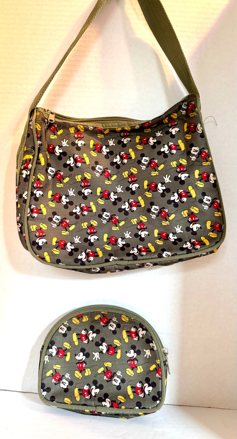May include: A gray shoulder bag and matching cosmetic bag feature a repeating pattern of Mickey Mouse in red, black, yellow, and white. The bags have olive green trim and straps, with zippered closures.