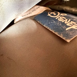 May include: Close-up of a tan fabric with a blue Disney tag. The tag is worn and has the Disney logo in a gold color. A white tag with text is partially visible.