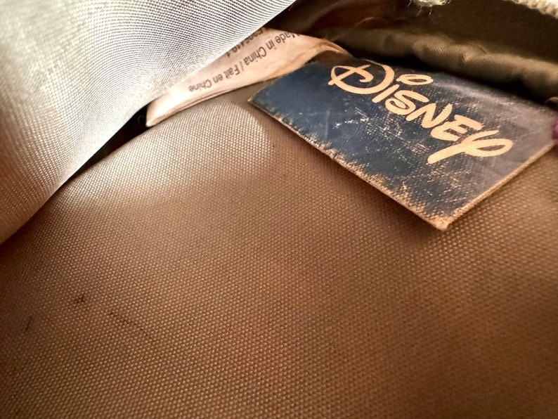 May include: Close-up of a tan fabric with a blue Disney tag. The tag is worn and has the Disney logo in a gold color. A white tag with text is partially visible.