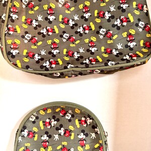 May include: Two-piece set featuring a large tote bag and a smaller, half-moon shaped cosmetic bag. Both are olive green with a repeating pattern of Mickey Mouse in red, black, yellow, and white. The bags have olive green trim and zippers.