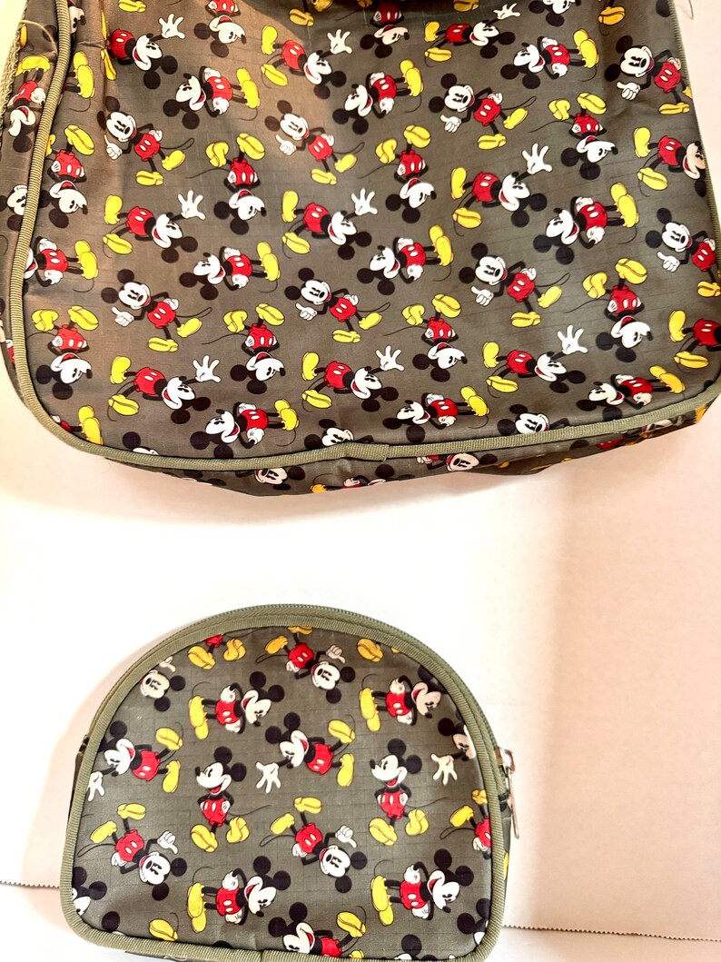 May include: Two-piece set featuring a large tote bag and a smaller, half-moon shaped cosmetic bag. Both are olive green with a repeating pattern of Mickey Mouse in red, black, yellow, and white. The bags have olive green trim and zippers.