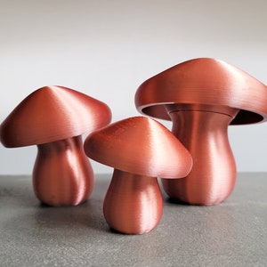 Peut inclure: Trois objets décoratifs en forme de champignon de couleur cuivre, de tailles différentes. Le plus grand champignon est le plus haut et a un sommet large et plat. Les deux autres champignons sont plus petits et ont des sommets plus arrondis.