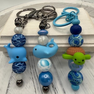 May include: Three beaded keychains with ocean themes. Each keychain features a variety of blue beads, a whale or turtle charm, and a silver-tone keyring. The keychains are displayed on a white wooden surface.