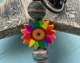 May include: A keychain with a black metal ring and clasp. The keychain features a colorful sunflower charm, patterned beads in teal, orange, and white, and a dark gray bead. The sunflower has petals in various colors.