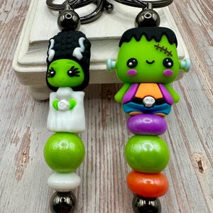 May include: Two Halloween-themed keychains. One features a green Bride of Frankenstein figure, the other a green Frankenstein's monster. Both have black keyrings and are made of colorful beads and charms.