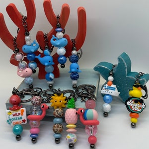 May include: A collection of beaded keychains with ocean-themed charms. Keychains feature charms like whales, flamingos, a pineapple, a sun, a cruise ship, and a duck. The keychains have various colored beads and metal hardware.