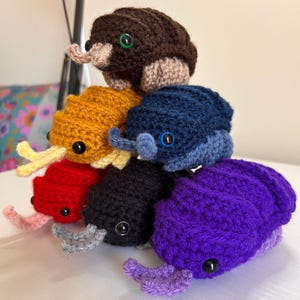 Może przedstawiać: Stos sześciu szydełkowych stworzeń przypominających robaki w różnych kolorach: brązowym, żółtym, niebieskim, czerwonym, czarnym i fioletowym. Każdy ma guziczkowe oczy, czułki i trąbkowaty pysk. Ręcznie robione zabawki są ułożone jedna na drugiej.