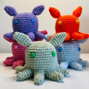 Puede incluir: Una colección de juguetes de pulpo hechos a mano con ganchillo en varios colores. Los juguetes tienen ojos de botón y pequeños tentáculos. Los colores incluyen morado, naranja, rosa, azul claro y verde. Los juguetes tienen un diseño caprichoso y juguetón.