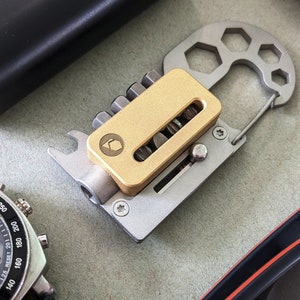 Puede incluir: Un llavero multiherramienta plateado con un porta-puntas de color dorado. La herramienta tiene un abridor de botellas, una llave hexagonal y un bucle para sujetarlo a un llavero.