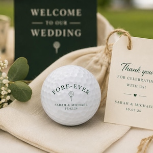 May include: A white golf ball printed with the text FORE-EVER, a golf tee icon, and the names Sarah & Michael with the date 10.02.26. Beside it is a beige fabric pouch and a cream-colored gift tag reading Thank you for celebrating with us!