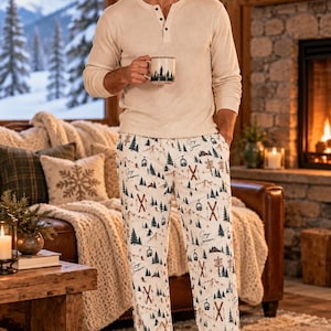 May include: A long-sleeve cream-colored henley shirt with a three-button placket and white pajama pants featuring a repeating print of mountains, pine trees, ski lifts, and the text Cozy Season, paired with a mug showing a forest silhouette.