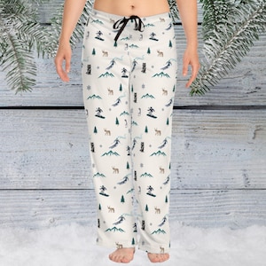 May include: Cream-coloured pajama trousers featuring a repeating pattern of skiers, deer, trees, and mountains. The trousers have a drawstring waist and a relaxed fit. The design includes blue, green, and brown elements.