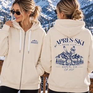 May include: A cream-coloured zip-up hoodie featuring a small mountain graphic and the word BRIDE on the front. The back displays a large blue graphic with skis, a martini glass, mountains, trees, and the text APRÈS-SKI BRIDE CLUB ASPEN, CO Est. 2027.