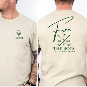 Könnte beinhalten: Beiges T-Shirt mit grünen Golfmotiven. Auf der Vorderseite befindet sich ein Golfball und das Wort "GROOM". Auf der Rückseite steht das Wort "Fore" in einer stilisierten Schriftart, zusammen mit Golfschläger- und Flaggen-Symbolen und dem Text "THE BOYS".