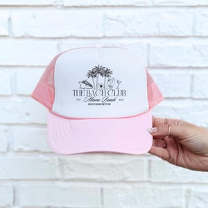 Puede incluir: Gorra de camionero rosa y blanca con la parte trasera de malla. La parte delantera de la gorra presenta el texto "THE BACH CLUB Miami Beach 2025" con ilustraciones de palmeras, un crucero y una copa de cóctel.