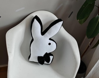 Peut inclure: Un coussin en forme de lapin blanc en peluche avec un contour noir et un nœud papillon noir est posé sur une chaise blanche avec des pieds en bois. La chaise est devant un mur gris avec des rayures grises et blanches.