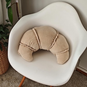 Op de afbeelding: Een beige croissantvormig kussen ligt op een witte stoel met houten poten. De stoel staat op een lichtbruin shagtapijt.